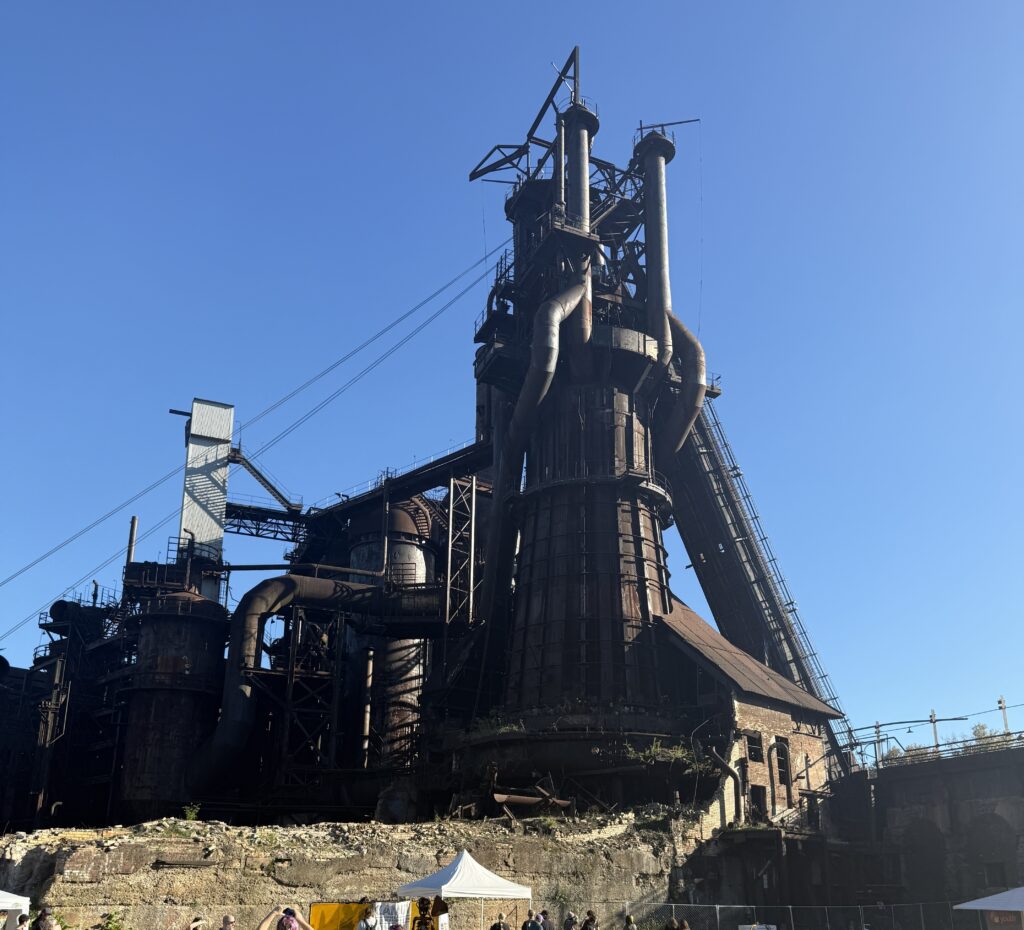 Historic Carrie Blast Furnaces rising against a clear blue sky, with festival tents and visitors below.