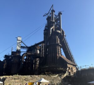 Historic Carrie Blast Furnaces rising against a clear blue sky, with festival tents and visitors below.